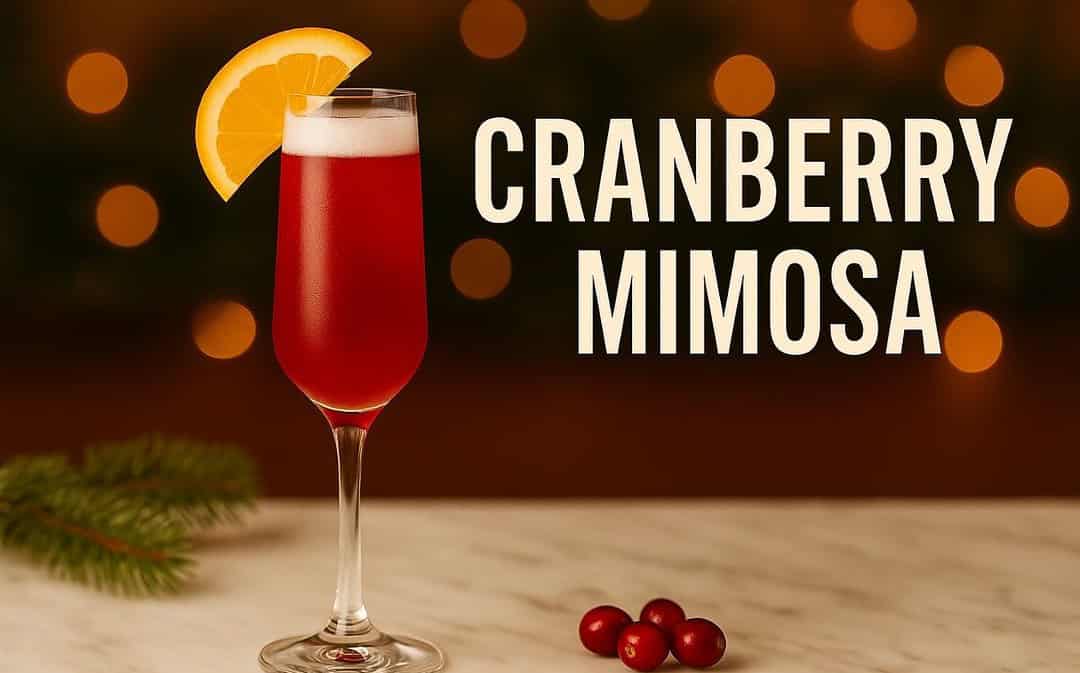 Cranberry Mimosa cocktail in a champagne flute with a bright red hue, orange slice garnish, and cranberries on marble with soft golden Christmas lights in the background.