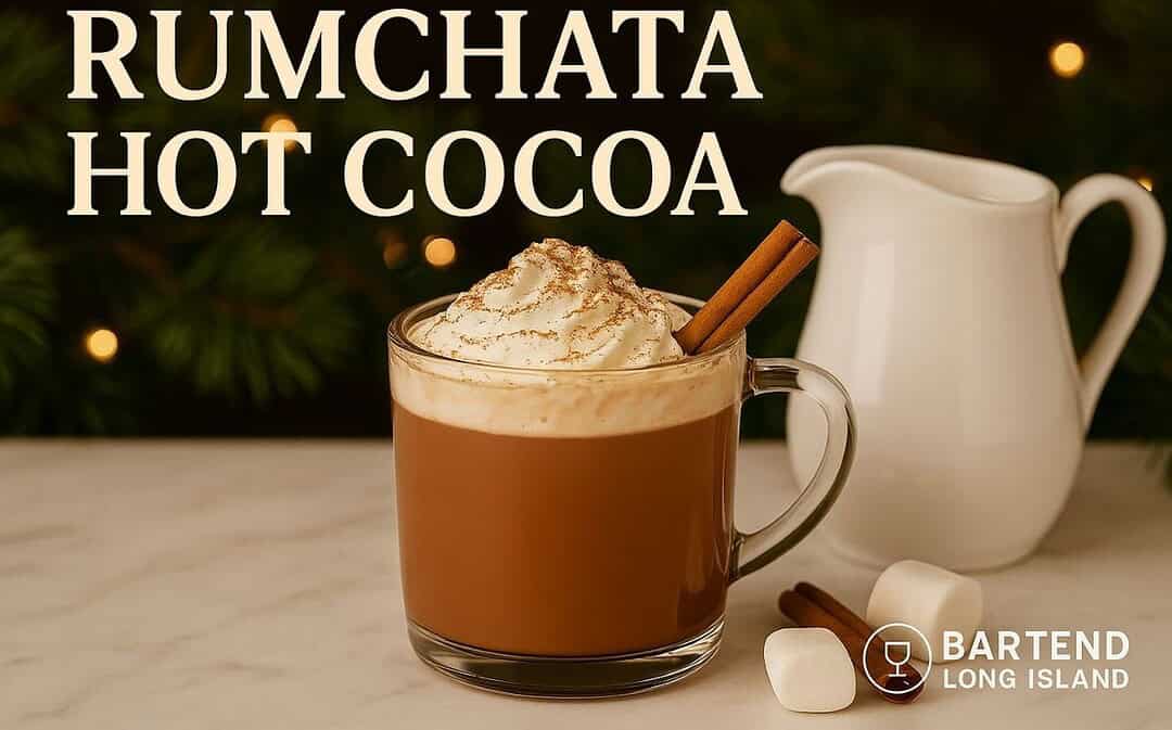 RumChata Hot Cocoa in a glass mug topped with whipped cream, cinnamon, and a cinnamon stick, with marshmallows and a white pitcher on marble against warm golden holiday lights.