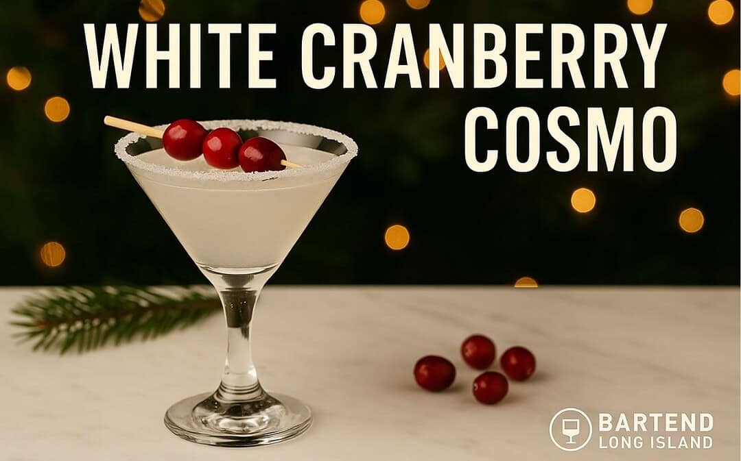 White Cranberry Cosmo cocktail in a martini glass with a sugar rim and cranberries on a cocktail pick, set on marble with pine branches and warm golden lights in the background.