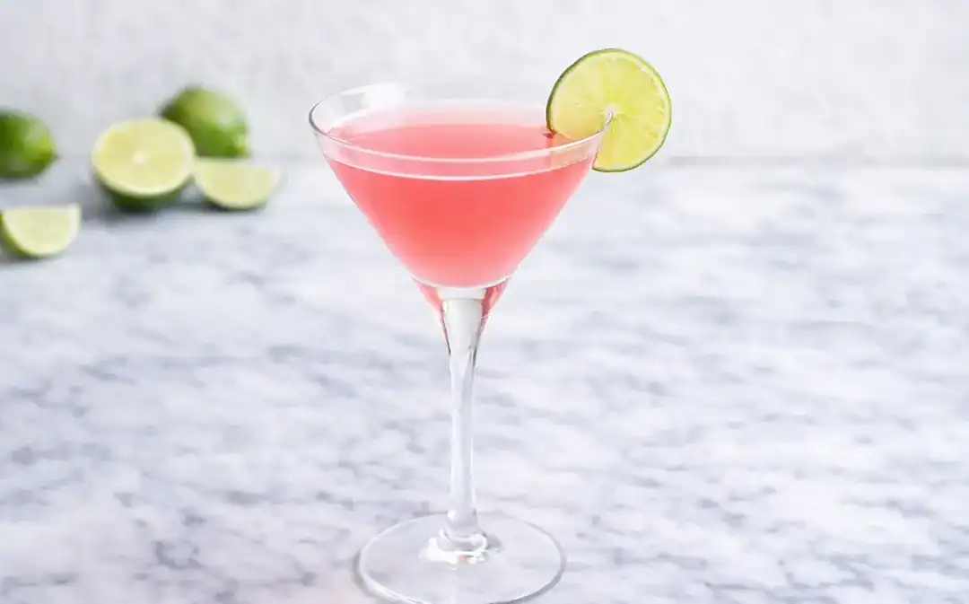 pink cosmo on marble counter top with lime wheel garnish and fresh cut limes in the background
