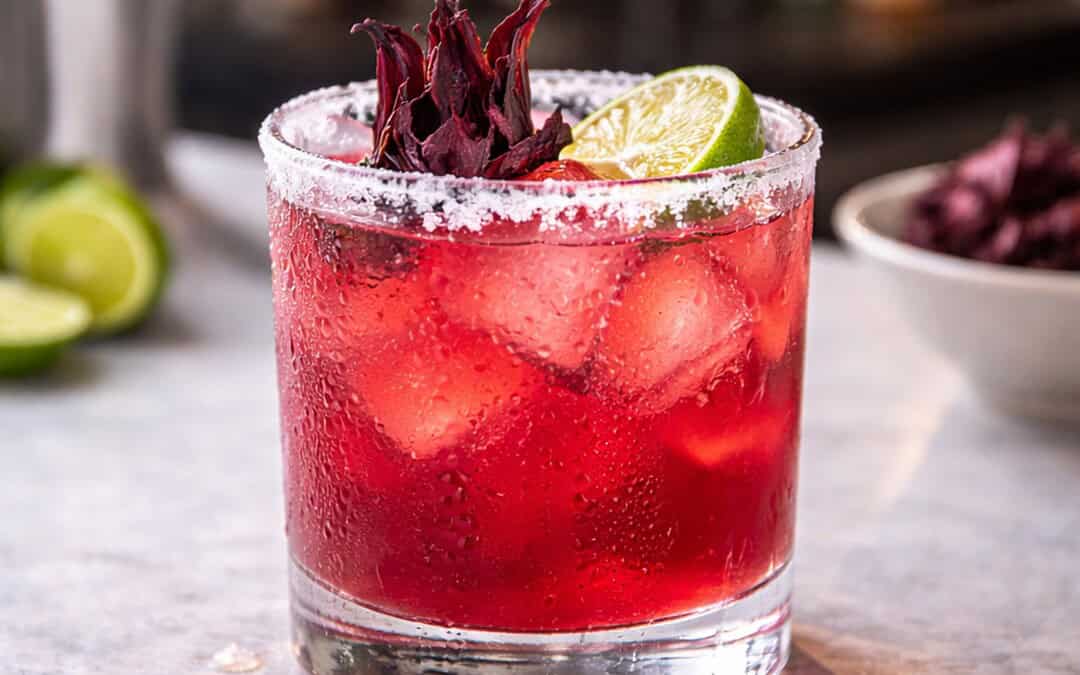 refreshing looking hibiscus margarita in a cold rocks glass with ice and a salt rim garnished with a fresh lime and hibiscus on a marble counter