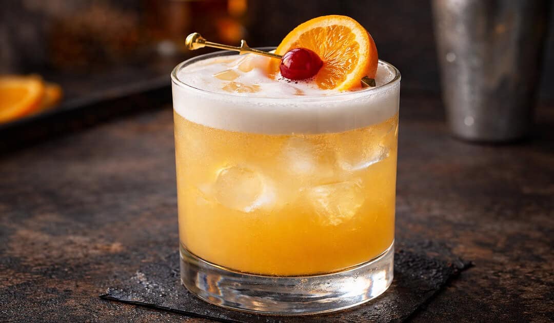 Classic whiskey sour cocktail with egg white foam, bourbon, lemon juice, and cherry orange garnish in a rocks glass