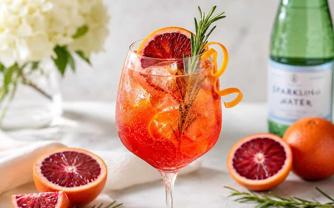 A blood orange spritz mocktail in elegant glassware with blood orange, rosemary, bubbles, citrus garnish, and ice.