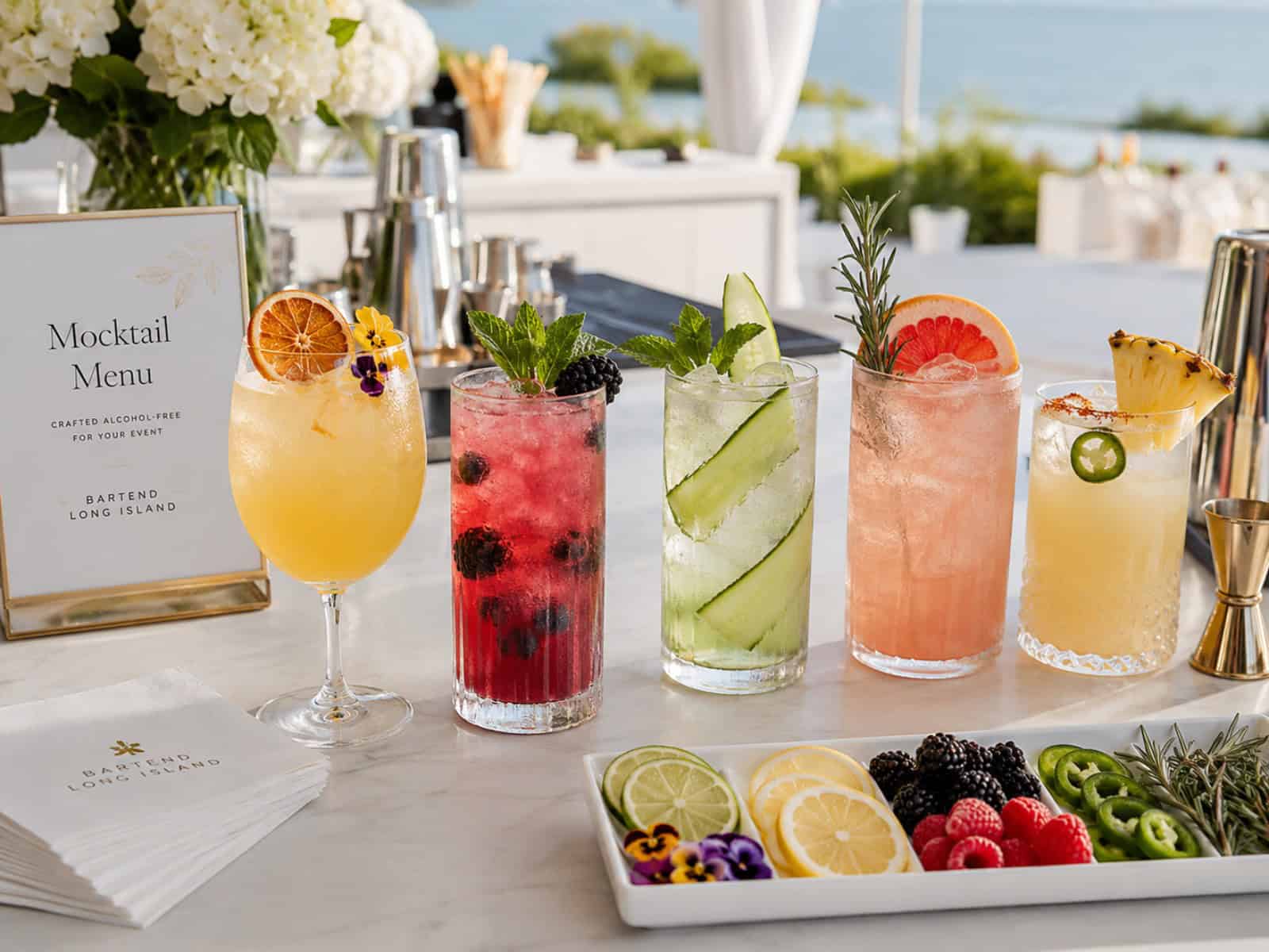 A private event mocktail bar setup with colorful alcohol-free drinks, fresh garnishes, citrus, herbs, bar tools, and elegant glassware.