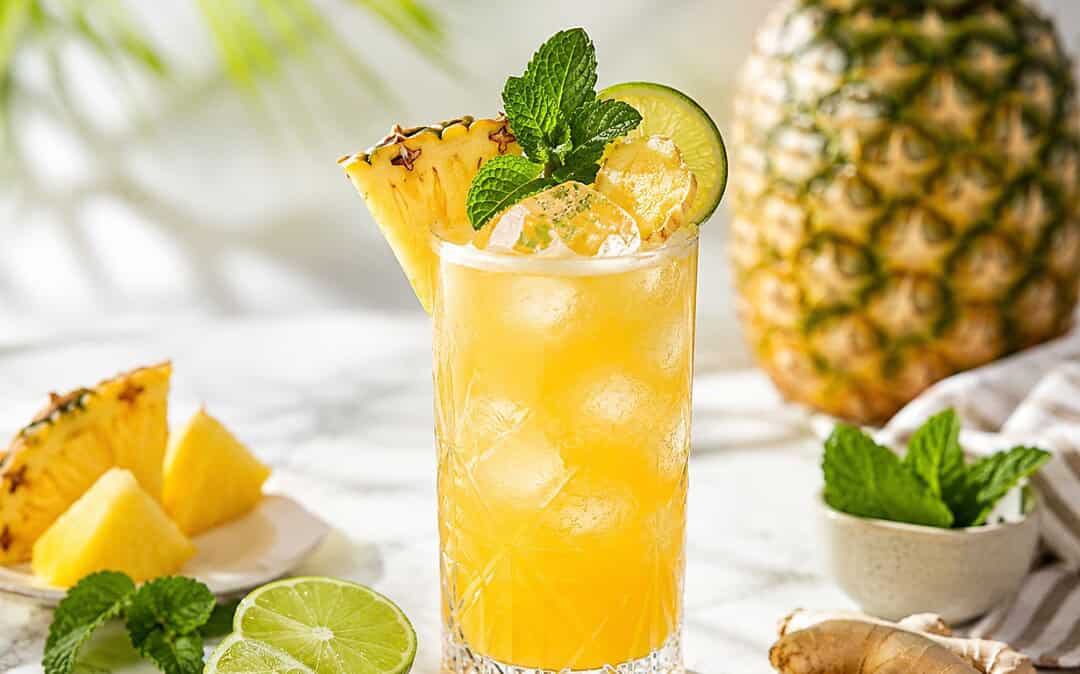 A pineapple ginger mocktail in elegant glassware with pineapple, lime, mint, fresh ginger, bubbles, and ice.